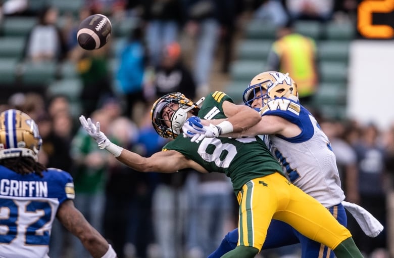 A football player leans toward a football in mid-air as a player on the opposing team tackles them. 