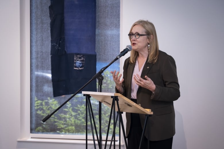 A white woman with round glasses speaks at a podium.