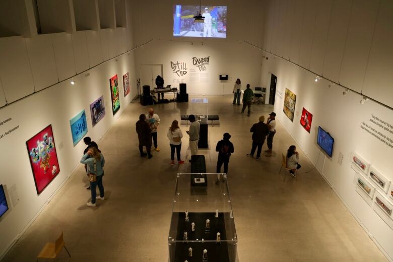 The view from above of an art gallery where people mingle. There's a video screen on one wall. 