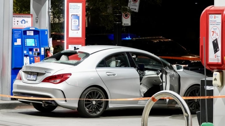 A silver car parked in a gas station. It's back window is broken. 