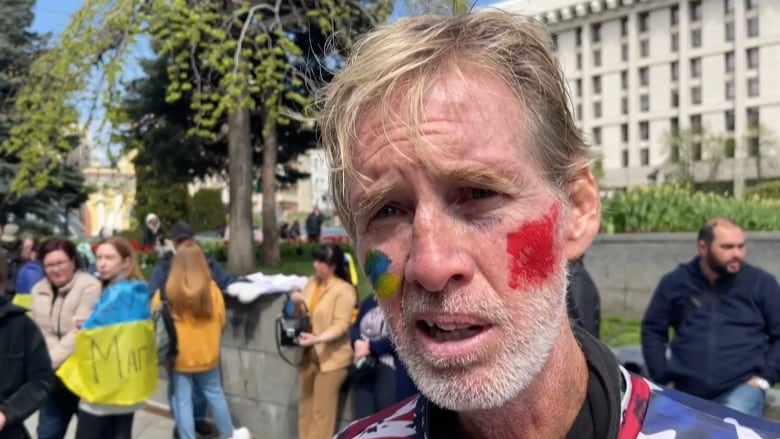 A bearded light-haired man with red and blue face paint on is shown in a closeup in an outdoor photo.