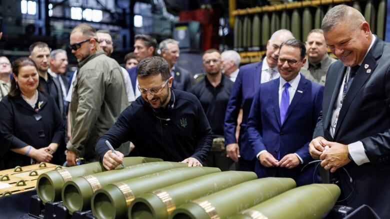 A smiling Zelenskyy writes on ammunition as men in suits look on.