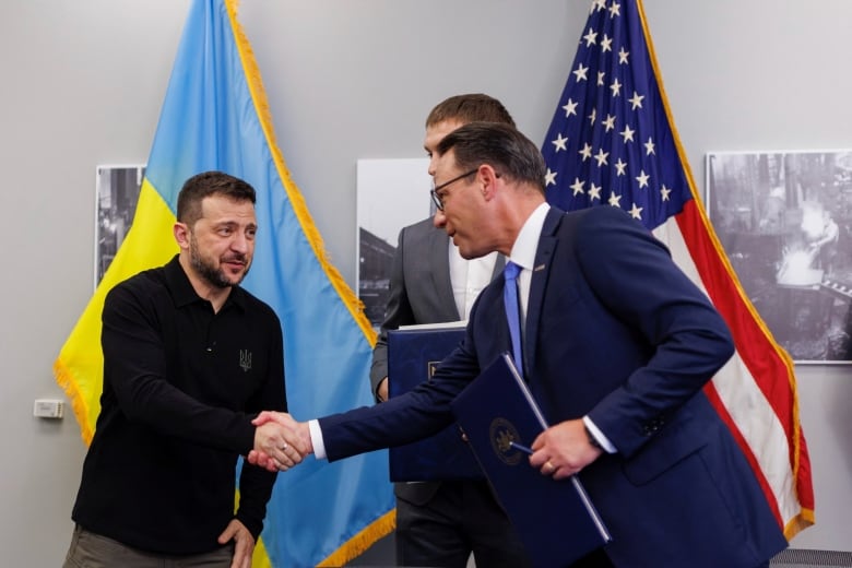 Zelenskyy smiling in a dark sweater and Shapiro in blue suit shake hands in front of the Ukraine and U.S. flags.