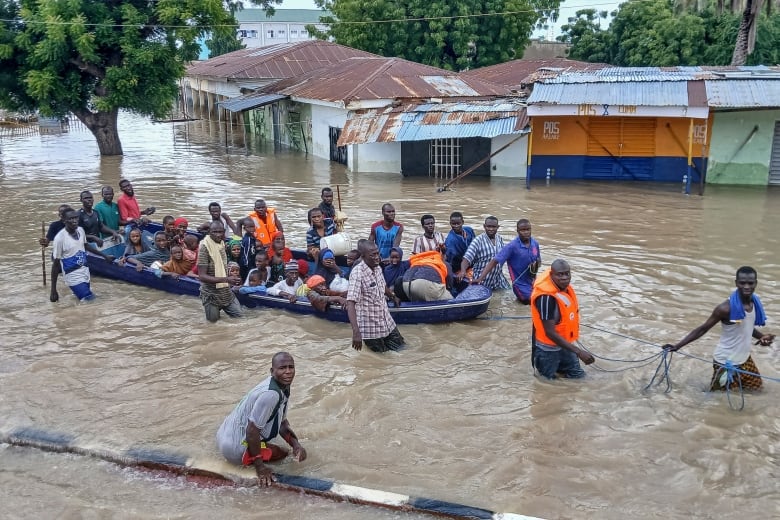 A low-slung boat in murky waters is filled with people. Others walk near it, hip-deep in water. 