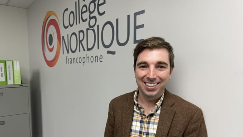 A man smiles at a camera with a wall that says Collge Nordique in the background. 