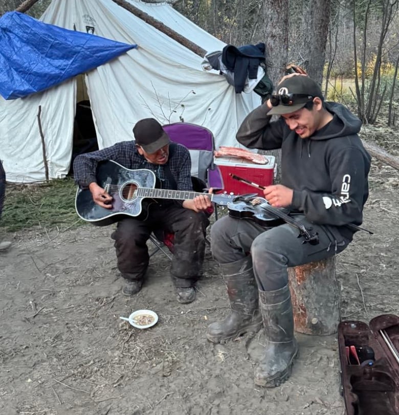 Two men sit at a campsite while one of them plays the guitar.