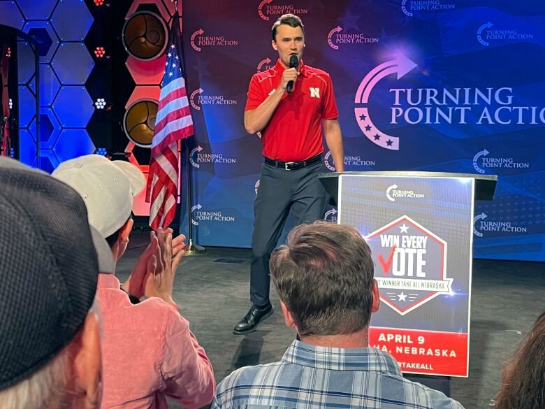 A cleanshaven young man in a polo shirt stands on a stage and speaks into a microphone. A placard that says 'win every vote' is shown as the backs of the heads of a few listeners are shown. 