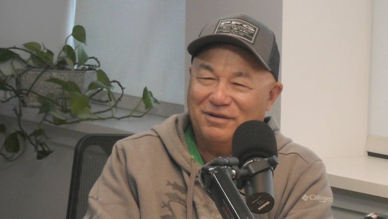 A man in a sage-green sweater and hat sits behind a microphone.