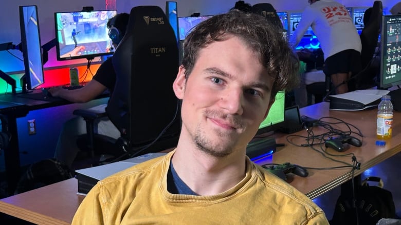 A white man with brown hair, wearing a yellow sweatshirt, sitting in a room with gaming consoles and computers. 