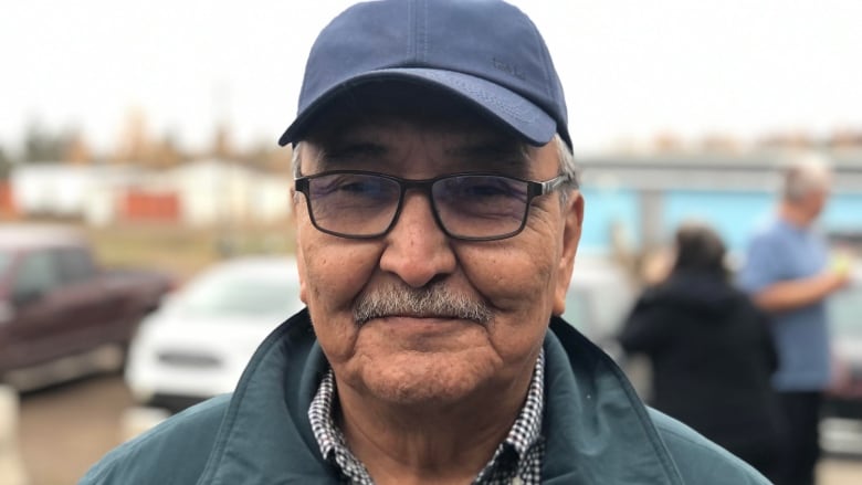 A man wearing glasses, a cap and a green collared jacket is seen in a close up headshot 