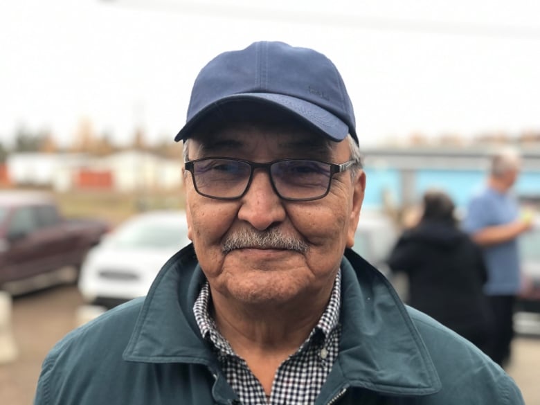 A man wearing glasses, a cap and a green collared jacket is seen in a close up headshot 