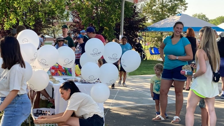 At a public park a woman bends down to tie a string of balloons together, there are a dozen more behind her. To her right two women are chatting while a small child watches the balloons.