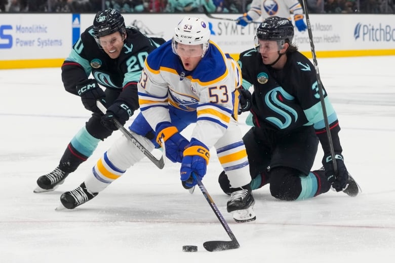 A male ice hockey player wearing number 53 skates with the puck as two opposing players pursue him during a game.