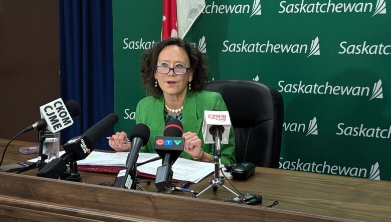 A woman in a green blazer sitting at a desk with multiple microphones in front of her