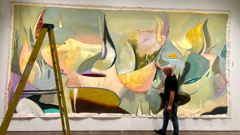 Scott Everingham, standing in front of his painting presented at the gala. The painting is a piece of abstract art featuring curves in the colours of green, pink, purple and yellow. Next to him is a large yellow ladder.  