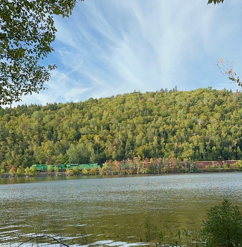 Across a lake, a train goes around the base of a wooded hill.