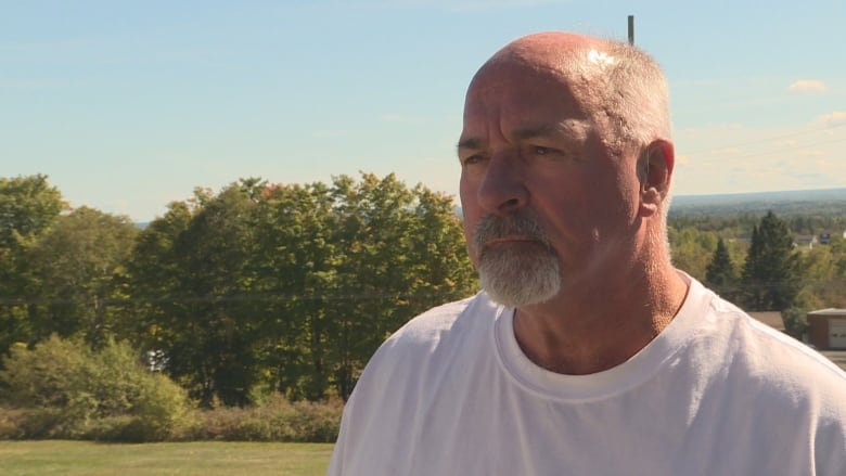 A man in a white tshirt with a bald head and grey moustache and beard. He's standing in front of the woods and looking to the left of the camera.