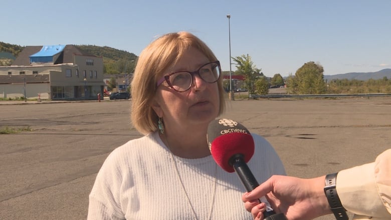 A woman with short blonde hair in a white sweater speaks to a reporter holding a microphone.