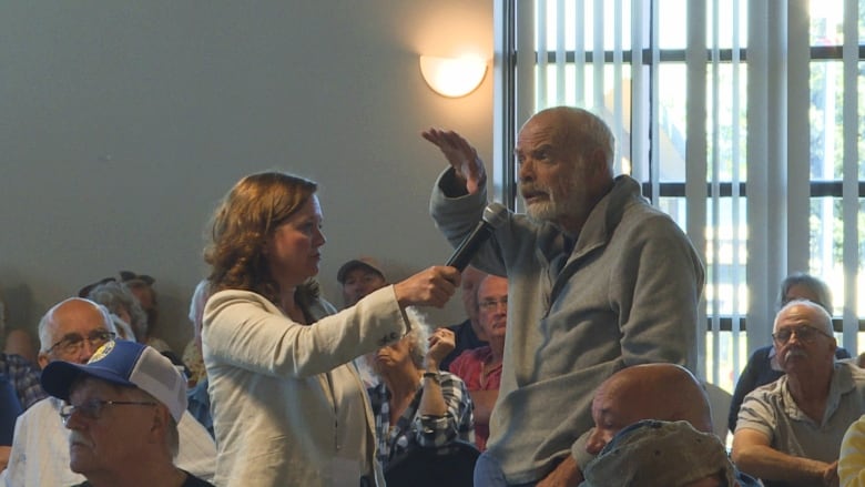 A man in a grey sweater speaks into a microphone being held by a moderator as other residents watch. He has an animated look on his face and his hand is raised.