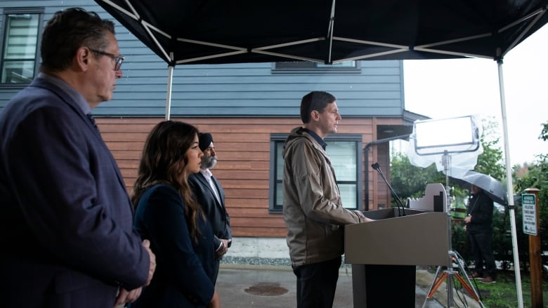 A side shot of a man standing at a podium with a few people standing behind him