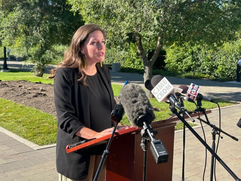 A wwoman in a black suit speaks in front of microphones with trees in the background. 