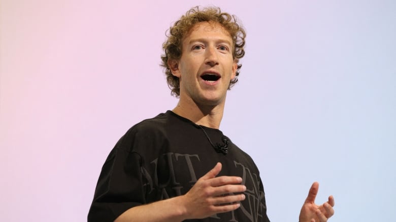 A man stands in front of a pastel background, clearly mid-speech, with raised arms. 