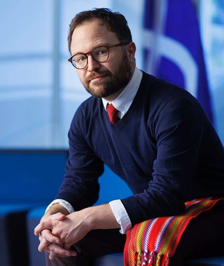 A man with round glasses looks into the camera sitting down with his arms on his knees.  