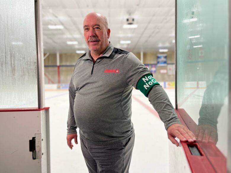 A man stands at the doors of a hockey rink with his arm extended on the door. His back is to the rink. He is wearing a green arm band.