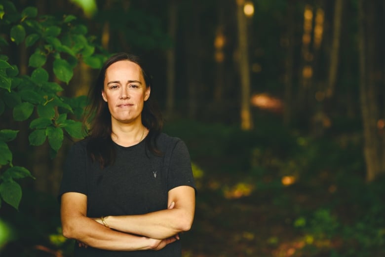 A woman with shoulder length brown hair wearing a black T-shirt with her arms crossed standing in front of green leaves.