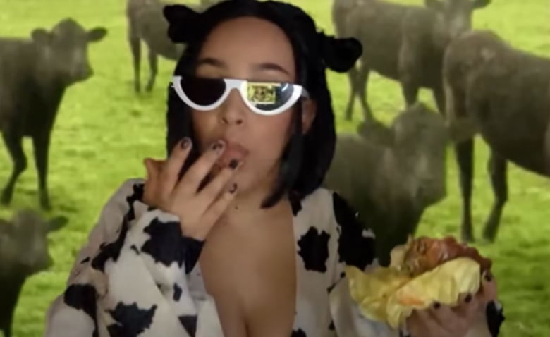 A woman in black and white half-moon sunglasses wearing a black and white spotted top puts her fingers to her mouth as cows mill around behind her on a grassy field.