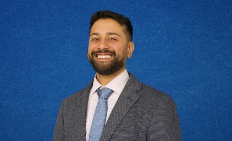 A man with black hair, black beard and tan skin smiles at the camera, wearing a grey suit and blue tie. 