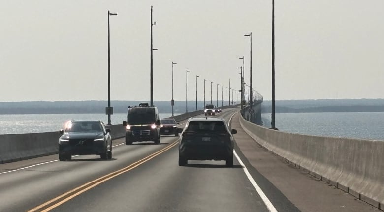 Cars and vans on a two-lane road on top of a curving bridge that is connected to land in the distance.