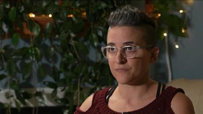 A young woman with glasses and short hair sits in her apartment. She is visible from the chest up.