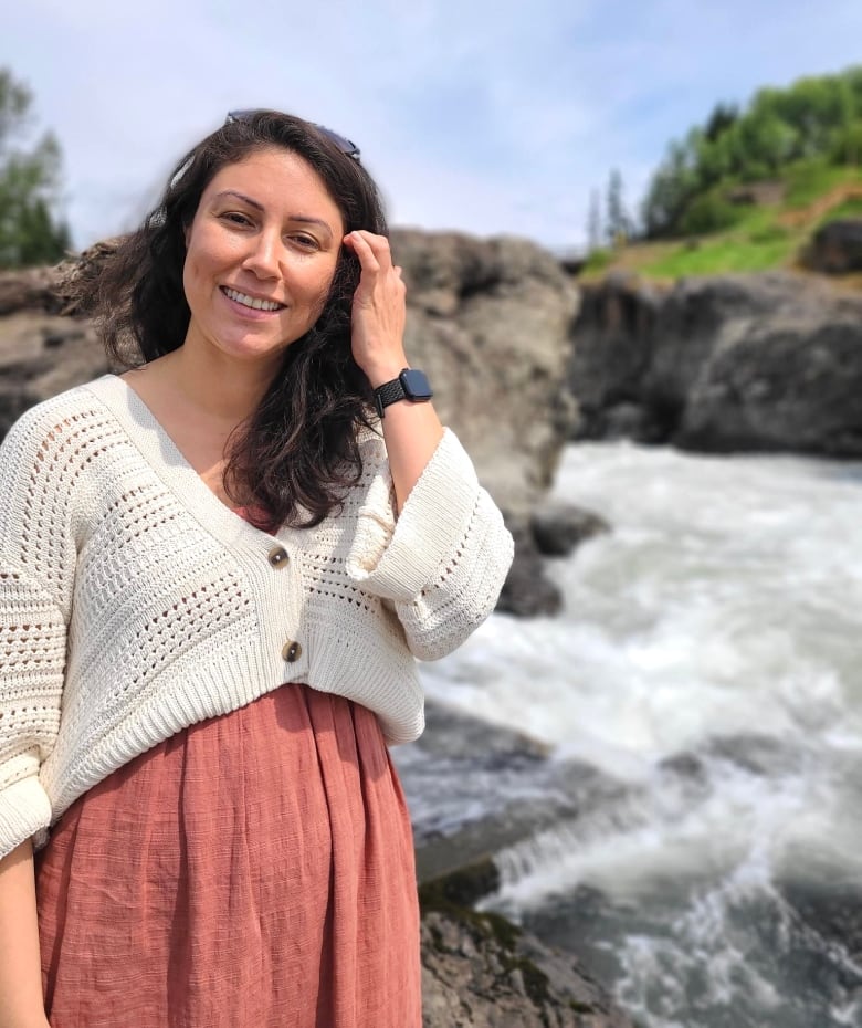 Christina smiles wearing a white sweater over an orange dress standing in front of a small waterfall. 