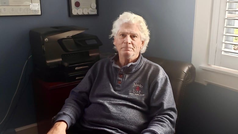 An older man with long wispy and curly hair has a neutral expression. He's sitting down with his arms on the chair. A printer and window are behind.