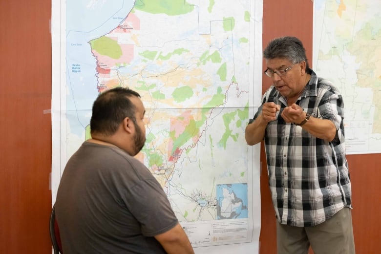 Two men stand talking and gesturing in front of a map of Cree territory in northern Quebec.