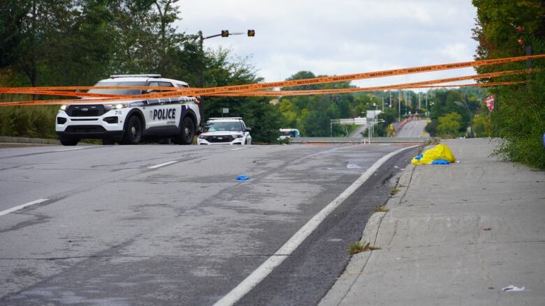 A police car is parked behind orange police tape that stretches across a city road on a cloudy day.