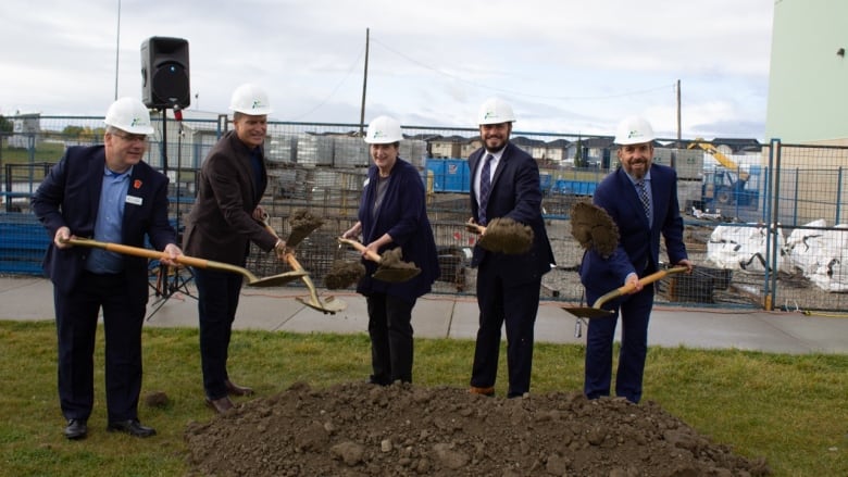 Five men and women wearing  white hard hats toss shovels full of dirt onto a lawn