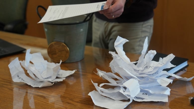 Some crumpled slips of paper are piled on a table with someone's hand visible in the background holding a piece of paper.