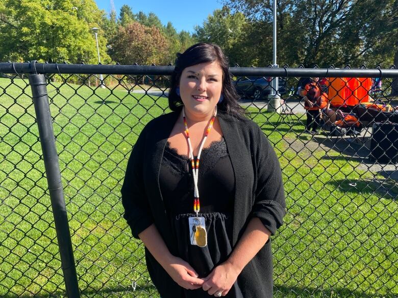 A woman in a black dress with a black sweater stands beside a fence with green grass and some people in orange shirts behind her.