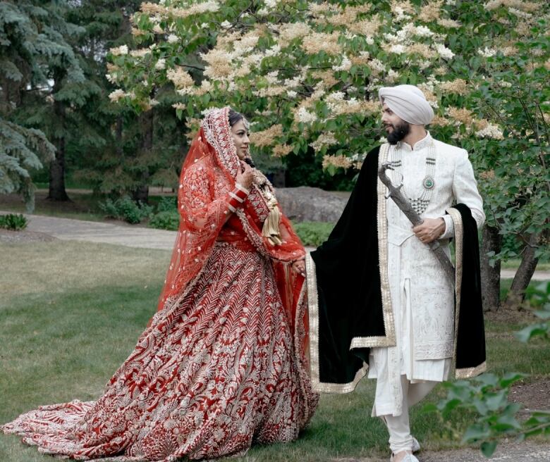 A woman in red and gold dress holds hands with a man in a white suit and turban.