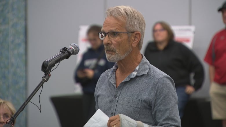 A man in a grey shirt stands at a mike ready to speak 