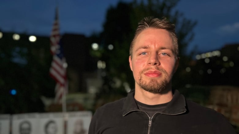 A man in a black top looks at the camera. He is standng outside a night, an American flag visible in the background.