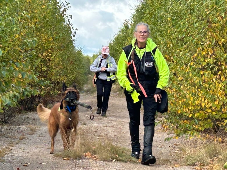 A woman walking along a tree-lined road with a Belgian Malinois beside her and a woman wearing a baseball hat a few feet behind them.