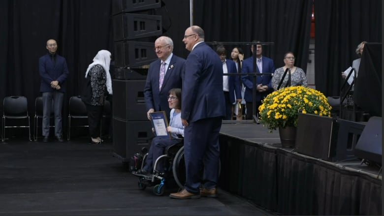 A woman in a wheelchair holds a framed award. Standing next to her are two men in suits. In the background a line of people wait to go on stage.