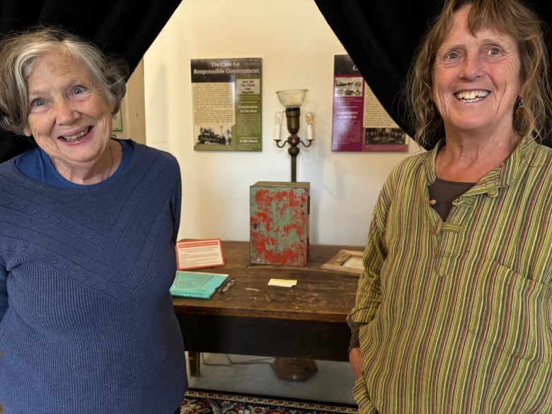 Two older woman, one in a blue sweater and the other in a striped green shirt, standing and smiling. Behind them is a table with a red box