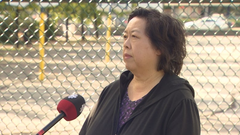 An East Asian woman wearing a black hoodie speaks in front of a wire mesh fence.