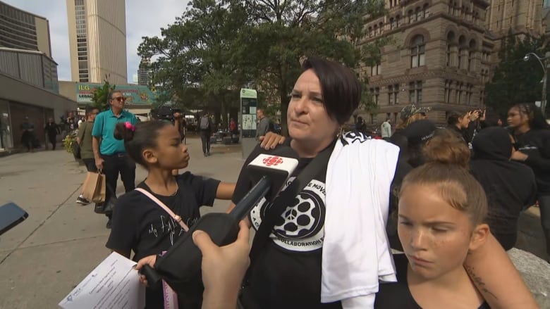 A middle age woman speaks with a reporter. She is outside Old Toronto City Hall, and has children on either side of her