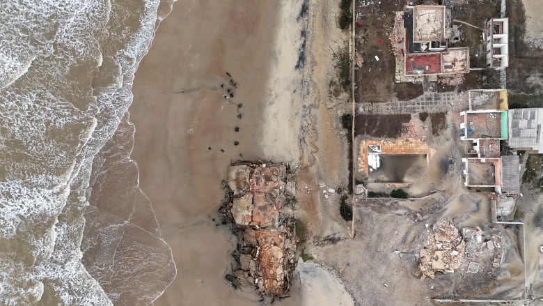 An aerial view pointing directly down at the ground shows a destroyed building on the beach, with a row of other houses behind it. 