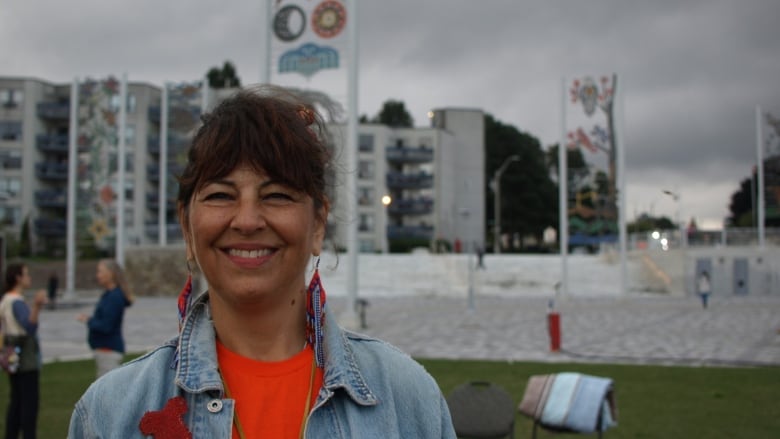 A person in an orange shirt and long earrings smiles. Some tall panels with glass art are in the background.